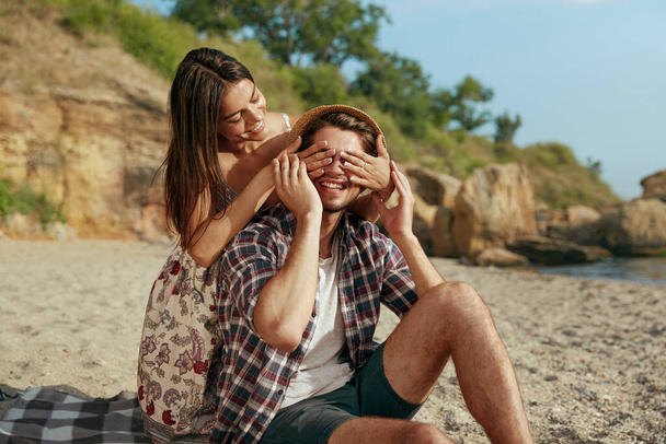 Happy girl closing eyes of her smiling boyfriend. Young european couple rest on sandy beach. Concept of romantic relationship and enjoying time together. Vacation on nature at summer. Sunny daytime - Фото, изображение