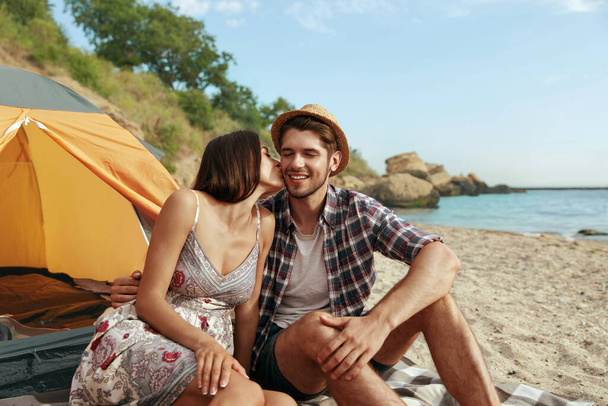 Beautiful girl kissing her pleased guy on sandy sea beach. Resting young european couple in love. Concept of romantic relationship and enjoying time together. Camping vacation at summertime. Sunny day - Zdjęcie, obraz