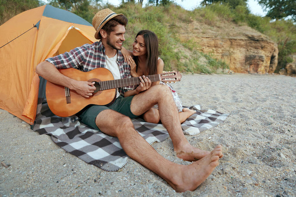 Happy guy play on guitar to beautiful smiling girl. Young european couple in love resting on sandy beach. Concept of romantic relationship and enjoying time together. Camping vacation at summertime - Foto, Bild