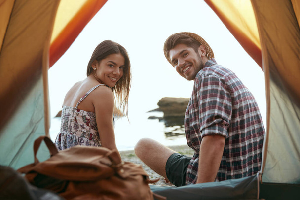 Young european couple resting on sandy sea beach. View from tent. Pleased guy and girl looking at camera. Concept of romantic relationship and enjoying time together. Camping vacation at summertime - Photo, image