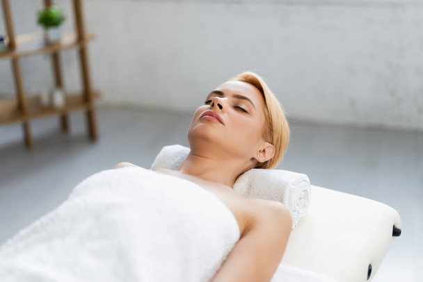 mulher loira com olhos fechados deitada no rolo da torre e mesa de massagem no centro de spa  - Foto, Imagem