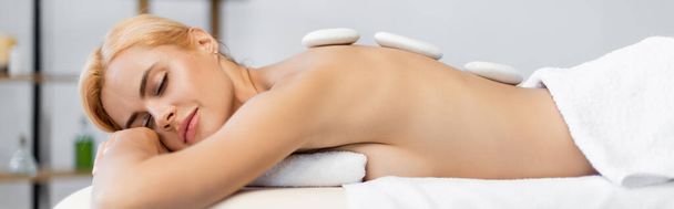 blond klient z zamkniętymi oczami otrzymujący gorący masaż kamienny w centrum spa, baner - Zdjęcie, obraz