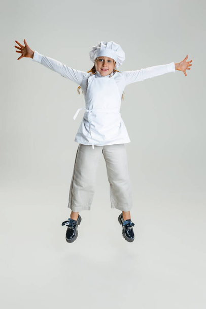 Studio shot of little cute girl in white cook uniform and huge chefs hat jumping isolated on white studio background. - Valokuva, kuva