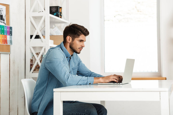 Młody brunetka człowiek pracuje z laptopem siedząc przy biurku w biurze wewnątrz - Zdjęcie, obraz