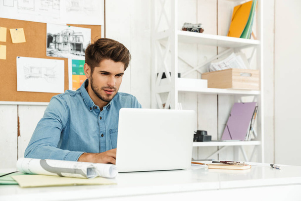 Νεαρός μελαχρινός άνδρας που εργάζεται με φορητό υπολογιστή, ενώ κάθεται στο γραφείο στο εσωτερικό - Φωτογραφία, εικόνα