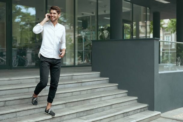 European businessman going on stairs and talking on mobile phone. Young man holding documents. Background of modern office building in city. Smiling bearded guy wear white shirt and jeans. Daytime - Fotó, kép