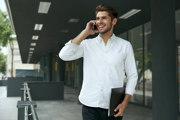 Pleased european businessman talking on mobile phone and holding documents. Young man standing on background of modern office building in city. Smiling bearded guy wear white shirt and jeans. Daytime - Fotoğraf, Görsel