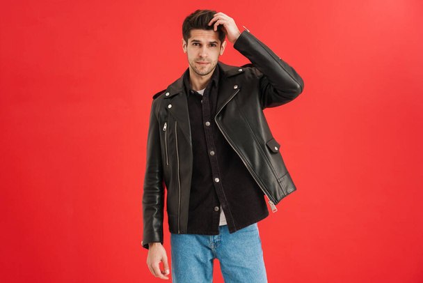 Young unshaven man wearing leather jacket looking at camera isolated over red wall - Valokuva, kuva