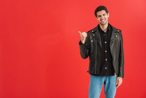 Young man wearing leather jacket pointing finger aside isolated over red wall - Фото, изображение