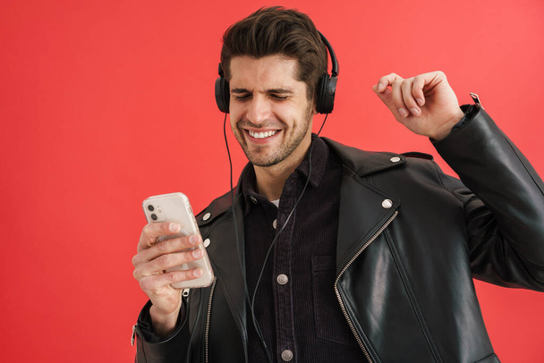 Young unshaven man dancing while using cellphone and headphones isolated over red wall - Fotó, kép