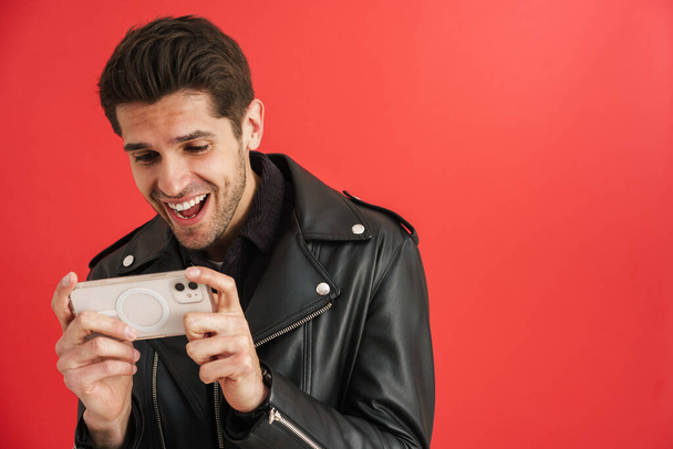 Young unshaven man laughing while using mobile phone isolated over red wall - Zdjęcie, obraz