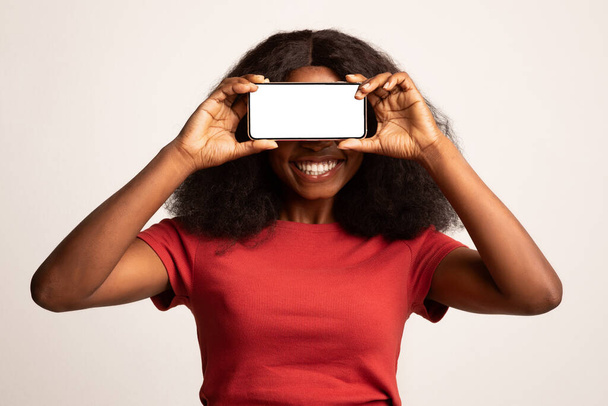 Mobile App. Cheerful Black Woman Covering Eyes With Blank Smartphone, Mockup - 写真・画像