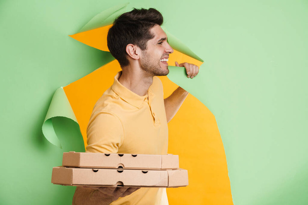 Smiling young casual white man looking out from a torn paper hole background holding takeaway pizza boxes looking aside - Photo, image