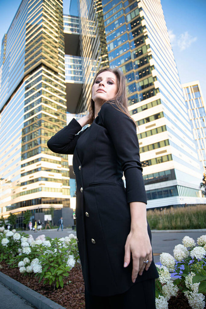 Young business woman in modern city of skyscapers in black suit with flower bed behind - Foto, immagini