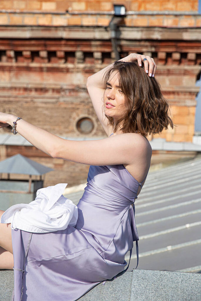 Brown haired young european woman sitting on the roof in lilac dress - Φωτογραφία, εικόνα
