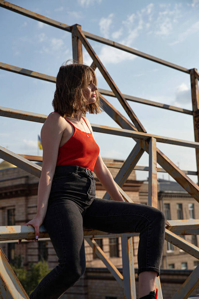 Young european woman in red top and dark jeans sitting on metal construction - Foto, Bild