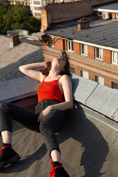 Young european woman in red top and dark jeans sitting on the roof with city view behind - Valokuva, kuva