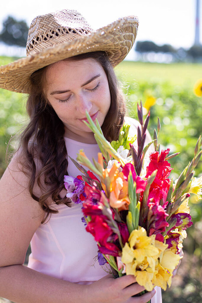 Portrait of white young woman with long hair in light dress in straw hat with bright bouquet of flowers in the countryside - Foto, afbeelding