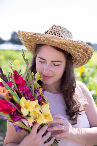 Portrait of white young woman with long hair in light dress in straw hat with bright bouquet of flowers in the countryside - Фото, изображение