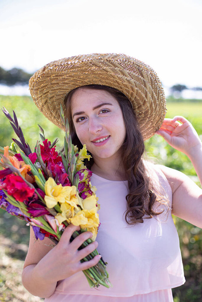 White young woman with long hair in light dress in straw hat with bright bouquet of flowers in the countryside - Фото, изображение