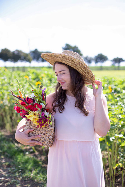 White young woman with long hair in light dress in straw hat with a basket of colorful flowers in the countryside - Foto, Bild