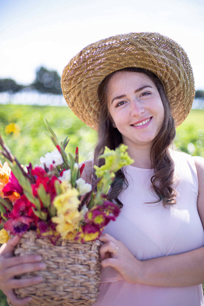 Portrait of smiling white young woman with long hair in light dress in straw hat with a basket of colorful flowers in the countryside - Foto, imagen