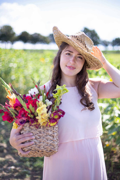 White young woman with long hair in light dress with a basket of colorful flowers in the countryside holding her straw hat by hand - Fotoğraf, Görsel