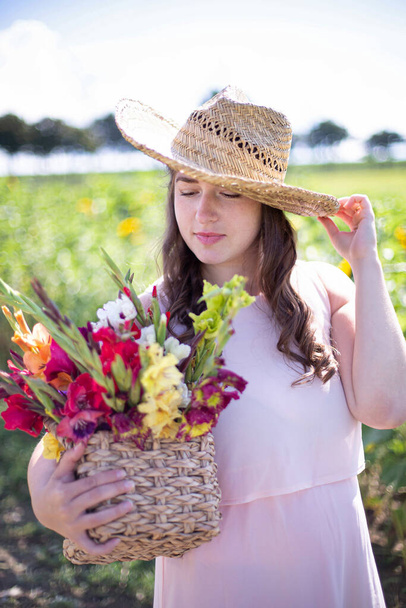 White young woman with long hair in light dress with a basket of colorful flowers in the countryside holding her straw hat by hand - Foto, afbeelding