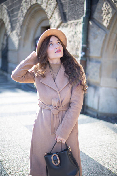 Cute pretty woman with curly hair in pastel pink coat with stone arches behind holding her hat - Fotó, kép
