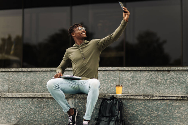Excited happy black african man, wearing glasses, takes selfie on mobile phone, wears casual fashion clothes on a walk in the city outdoors - Foto, Imagem