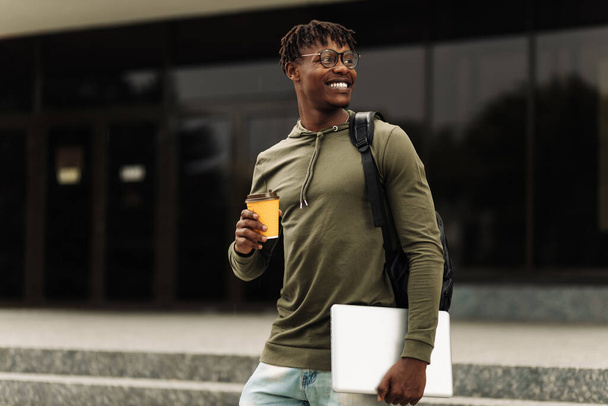 Stylish smiling African American man in glasses, drinking coffee in a cup and walking up the stairs in the city center with a laptop in his hands, stylish freelance people, with a laptop - Foto, Imagem