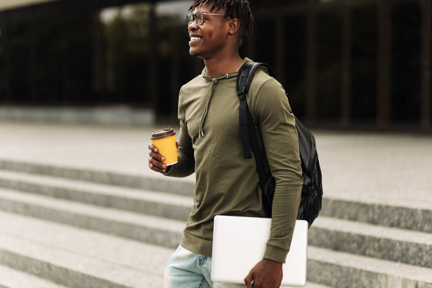 Stylish smiling African American man in glasses, drinking coffee in a cup and walking up the stairs in the city center with a laptop in his hands, stylish freelance people, with a laptop - Φωτογραφία, εικόνα