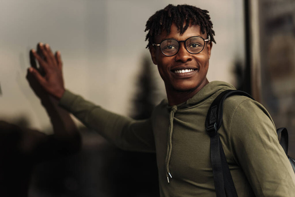 Happy handsome African American man, wearing glasses and with a black backpack, stands near a glass building smiling, looks into the frame on the street - Φωτογραφία, εικόνα