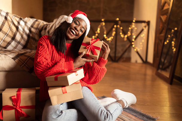 Święta i Nowy Rok. Radosny młody czarny kobieta w Santa kapelusz gospodarstwa prezenty świąteczne, siedzi na podłodze w domu - Zdjęcie, obraz