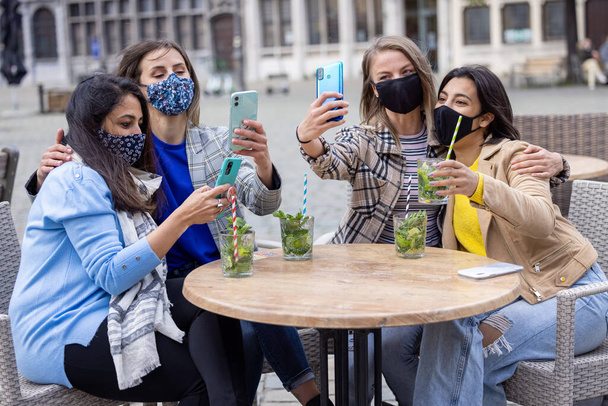 Amigos jovens multirraciais usando máscara facial enquanto tomam selfie com câmera de smartphone móvel enquanto bebem coquetéis em um terraço de um café na cidade durante o surto do vírus da corona - Foto, Imagem