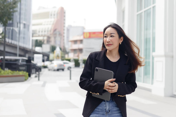 Beautiful asian businesswoman wear black suit holding tablet walking city street. woman freelancer lifestyle human urban. copy space use for text word. - Zdjęcie, obraz