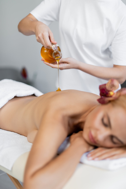 masażysta trzymając dzbanek podczas wylewania olejku do masażu na plecy rozmytego klienta - Zdjęcie, obraz