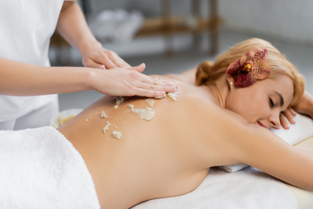 masajista aplicando exfoliante corporal en la parte posterior del cliente en la mesa de masaje - Foto, Imagen