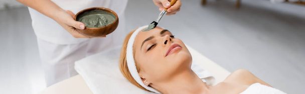 schoonheidsspecialiste aanbrengen klei masker op gezicht van vrouw liggend op massagetafel, banner - Foto, afbeelding