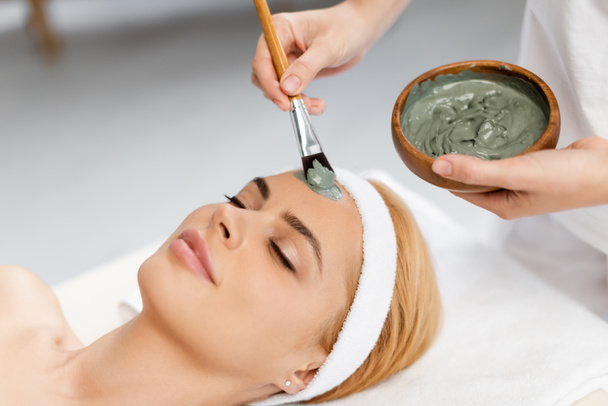 beautician holding bowl and applying clay mask on face of woman in spa center - Фото, зображення