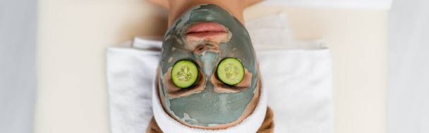 bovenaanzicht van vrouw met kleimasker en verse komkommerschijfjes op ogen liggend op massagetafel, spandoek - Foto, afbeelding