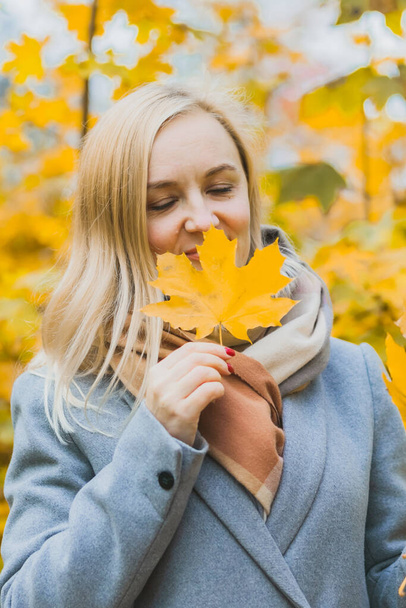 Een vrouw die een geel esdoornblad in haar handen snuift in een park tegen een achtergrond van bomen, geniet van de herfst en de natuur. Authentieke 40-jarige vrouw zonder retoucheren - Foto, afbeelding