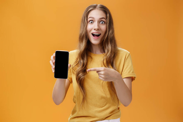 Girl surprised with awesome smartphone features. Portrait of delighted and amazed cute european slender woman with wavy natural hairstyle showing cellphone pointing at gadget screen at app - Photo, image