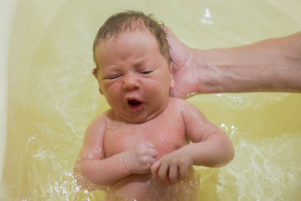 A newborn baby is bathing in a bath for the first time, his hand is holding his head, the baby is scared. - Fotografie, Obrázek