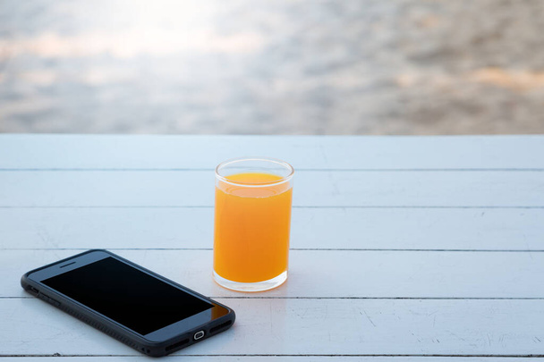 Sumo de laranja em vidro e telefone celular em fundo de mesa de madeira branca - Foto, Imagem