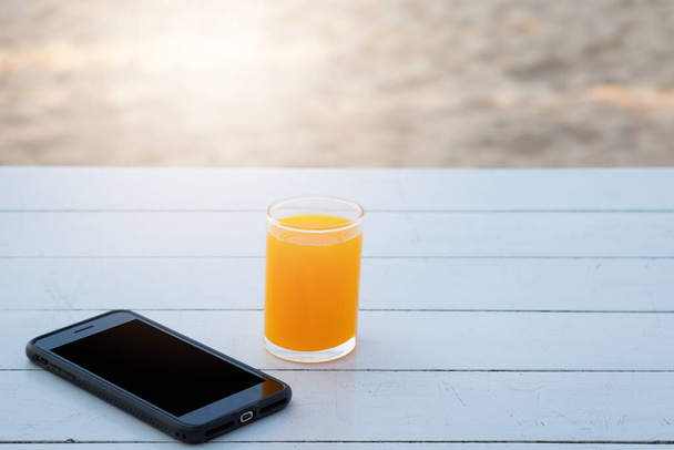 Апельсиновый сок в стекле и мобильный телефон на фоне белого деревянного стола - Фото, изображение
