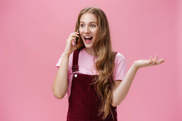 Portret popularnej wychodzącej i rozmownej młodej atrakcyjnej dziewczyny w kombinezonie machającej ręką podczas rozmowy na smartfonie rozbawiony i podekscytowany plotkowaniem z przyjacielem przez telefon komórkowy - Zdjęcie, obraz