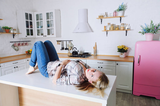 Young cute blonde woman 30+ in blue jeans and a shirt sitting and relaxing in her bright clean kitchen. Healthy happy lifestyle. Leisure. - Фото, изображение