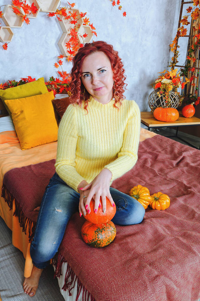 Beautiful redhead curly young woman in warm sweater sits in the bed at a cozy home, laughing, enjoying. Home interior with autumn decorations, orange leaves, flowers, pumpkins.  - Fotó, kép