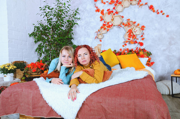 Twee mooie jonge vrouwen, zussen, vrienden in warme truien zitten in bed in een gezellig huis, lachend, genietend. Binnenhuis met herfstdecoraties, sinaasappelbladeren, bloemen, pompoenen.  - Foto, afbeelding